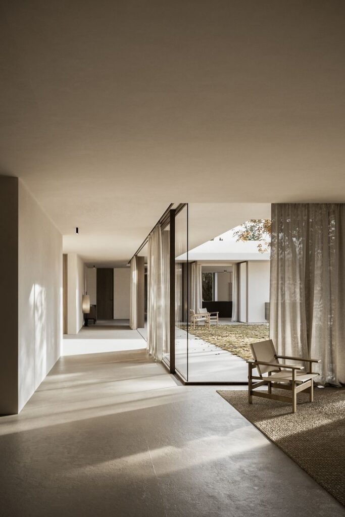 Modern, minimalist indoor space with large glass doors opening to a courtyard. Sunlit areas have beige flooring and walls, while wooden chairs and neutral curtains enhance the simple decor.