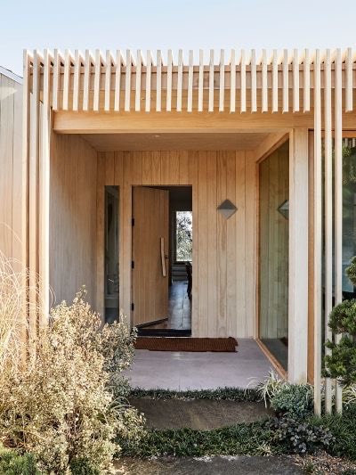 Modern home entrance with wooden cladding, vertical slats, and glass panels, surrounded by minimalist landscaping.