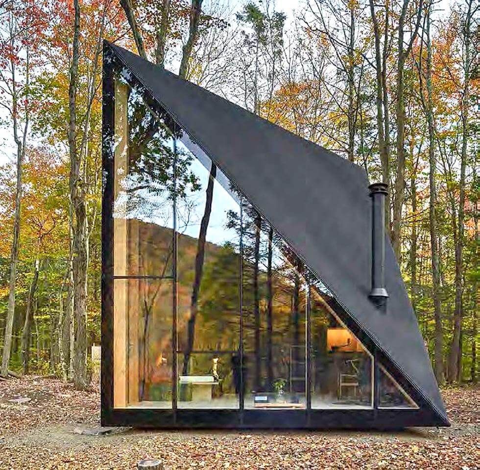 Modern A-frame cabin with large glass windows, nestled in an autumn forest setting, showcasing contemporary architecture.