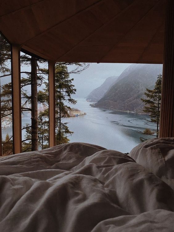Scenic mountain and lake view from a cozy bed in a cabin, framed by wooden beams and tall pine trees.