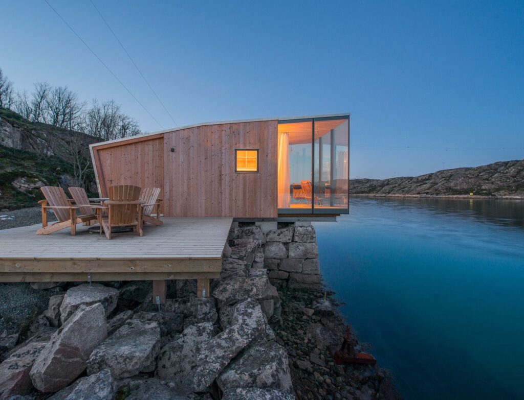 Modern wooden cabin with glass walls overlooking a serene lake, featuring an outdoor deck with wooden chairs at dusk.