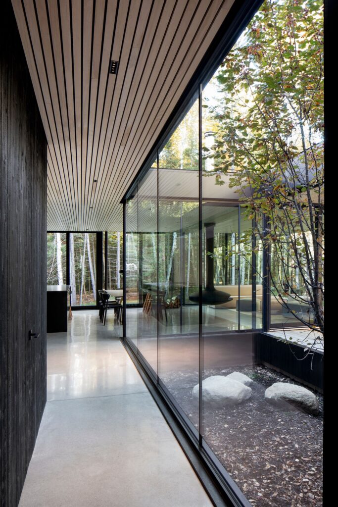 Modern interior with large floor-to-ceiling glass windows overlooking a garden area. The space features a minimalist design with wooden ceiling panels and polished concrete floors.