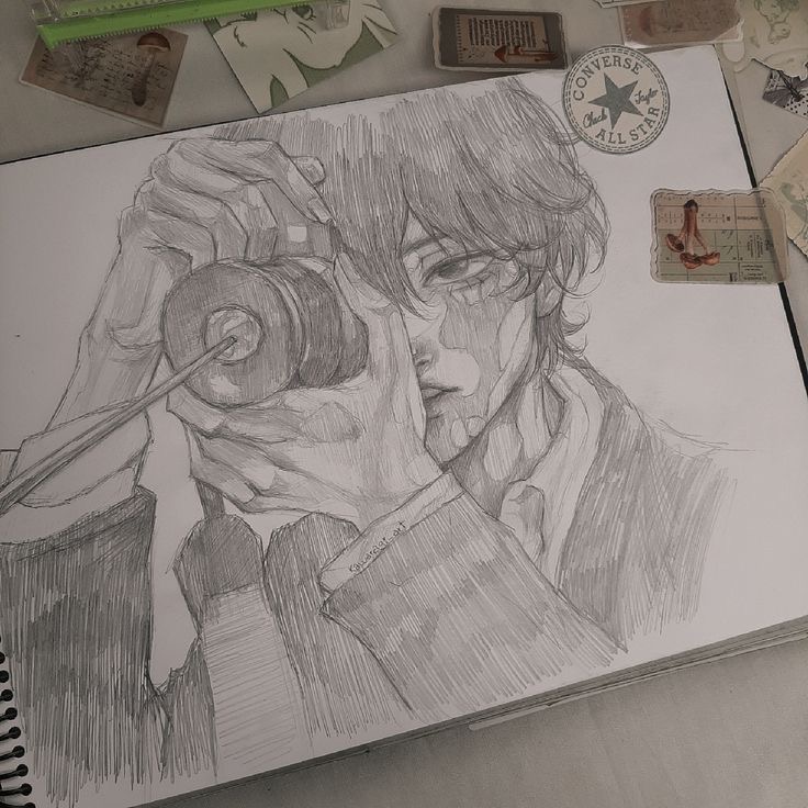 A detailed pencil sketch of a person holding a camera up to their eye, appearing to take a photo. The drawing lies on an open sketchbook amidst scattered paper items and a Converse emblem item.