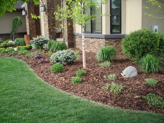 Beautifully landscaped front yard with mulch, green grass, various shrubs, and a small tree near a stone-accented house.