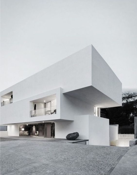A modern, minimalist white building with cantilevered upper floors and clean lines, featuring large windows and a rock set in front on a paved area.