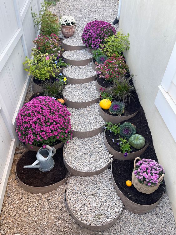 Beautiful garden pathway with circular pebble steps, colorful flowers, and pumpkins, bordered by white fence and wall.
