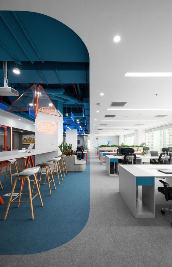 Modern office space with open workstations, lounge area, and vibrant decor featuring neon hello sign and blue accents.
