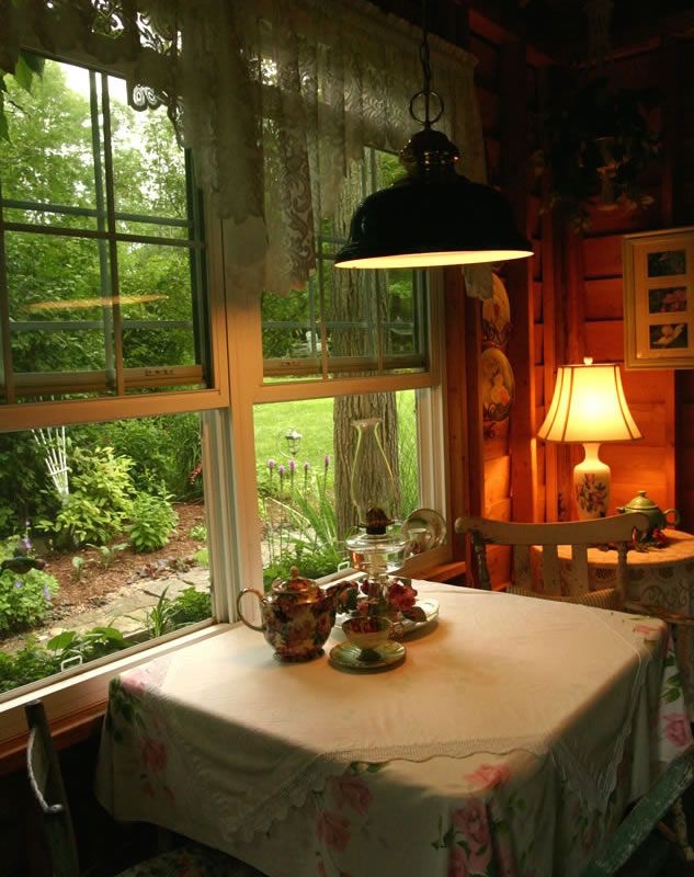 Cozy cottage table setup by a window with floral teapot, tiered tray, lace curtains, and garden view, under warm lighting.