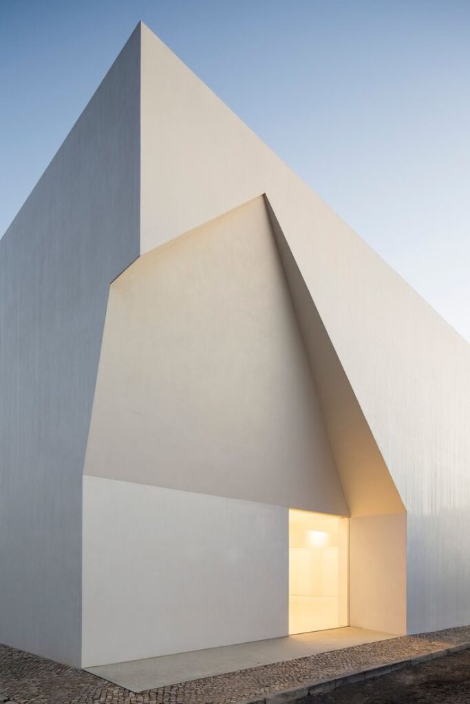 A minimalist building with sharp angles and a geometric design features a main entrance illuminated from within at dusk.