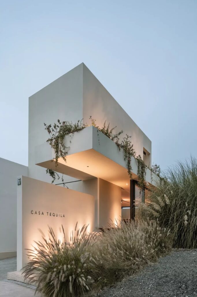 A modern, minimalist house named "Casa Tequila" with clean architectural lines, white exterior walls, and plants adorning the upper level and surrounding landscape.