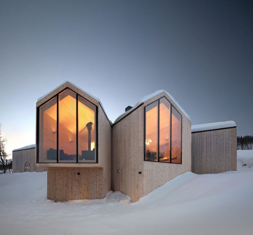 Modern cabin with large glass windows illuminated from within, surrounded by snow under a twilight sky.