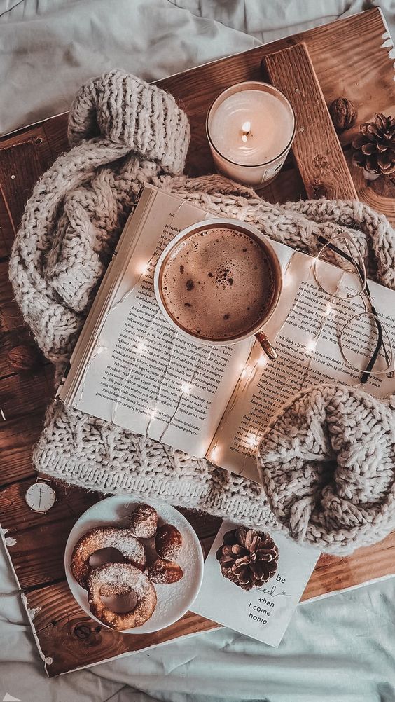 Cozy flat lay with an open book, hot coffee, knitted blanket, candle, glasses, and donuts on a wooden tray. Warm and inviting.