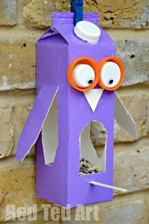 DIY bird feeder made from a painted milk carton resembling an owl, hanging on a brick wall, filled with bird seeds.