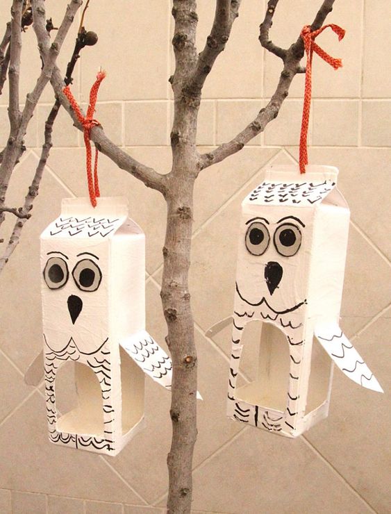 DIY owl bird feeders made from recycled milk cartons, painted white and black, hanging from tree branches with orange strings.