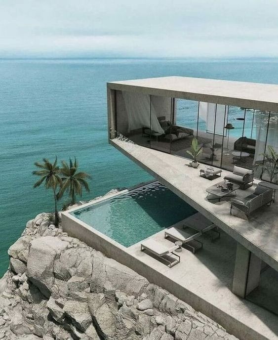 Modern cliffside house with floor-to-ceiling glass walls overlooks a body of water. The home features an infinity pool, palm trees, and outdoor lounging areas.
