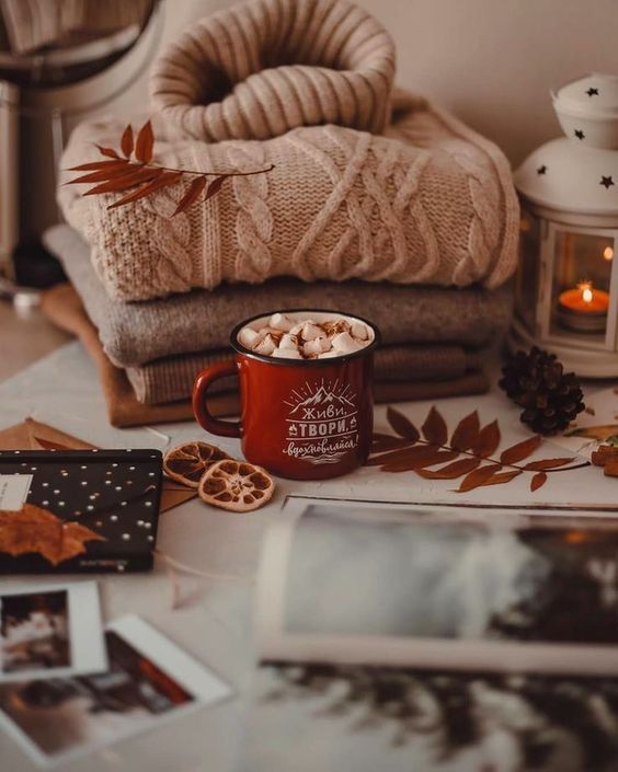 Cozy autumn setup with a red mug of hot cocoa, sweaters, dried leaves, and a candle lantern on a table.