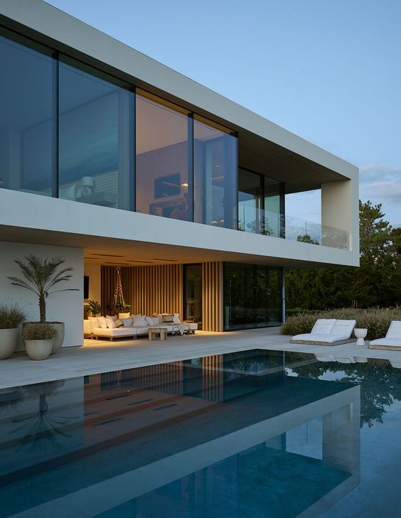 Modern luxury house with floor-to-ceiling windows, large outdoor pool, and stylish patio furniture at sunset.