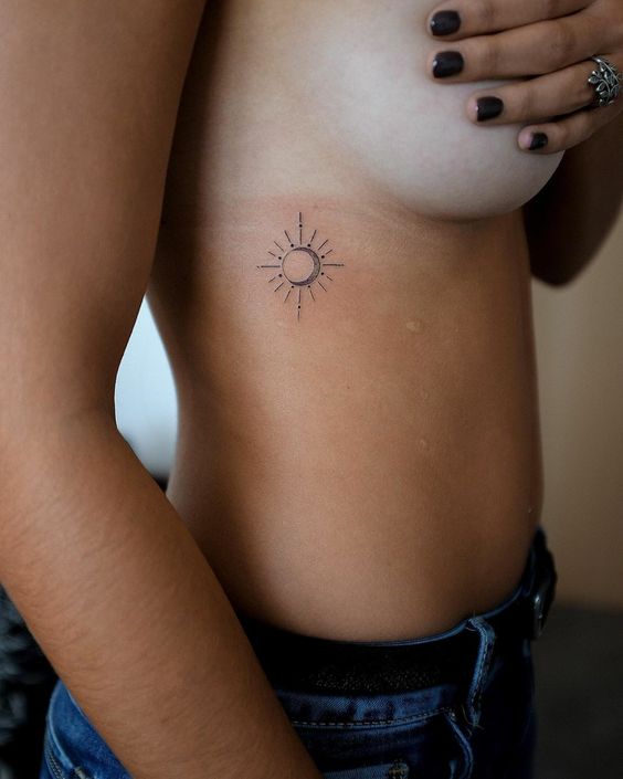 Minimalist sun tattoo on left ribcage, woman in blue jeans with dark nail polish. Elegant side body art design.