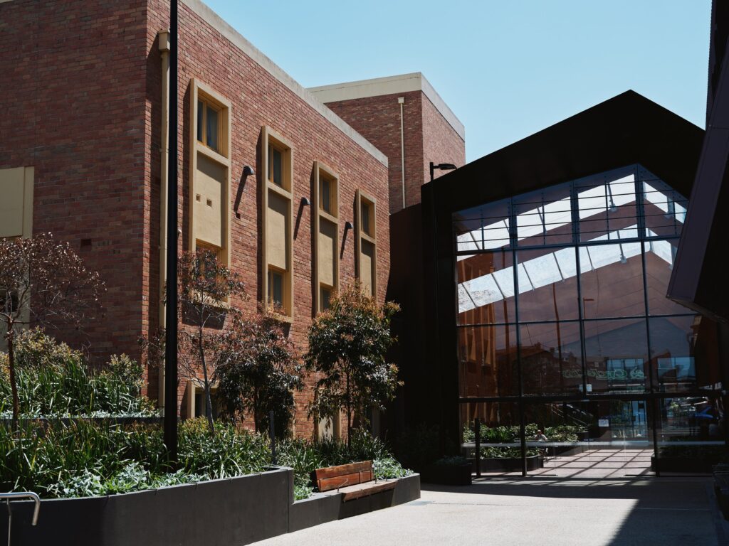 Modern building with brick walls and large glass windows reflecting the blue sky, surrounded by greenery and pathways.