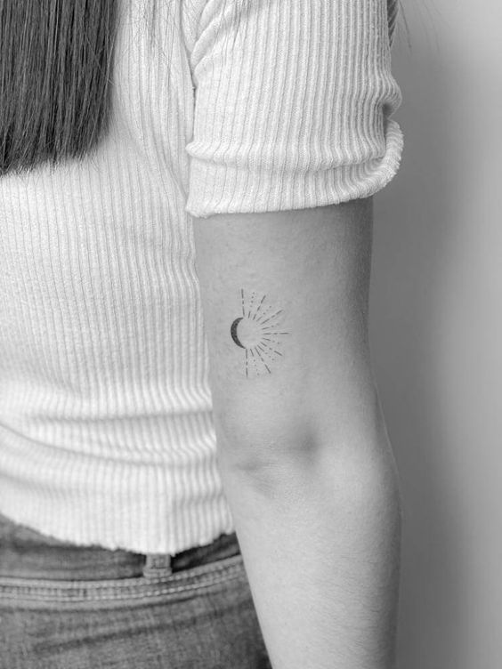 Minimalist crescent moon tattoo on a woman's upper arm, shown in black and white, wearing a ribbed white top.