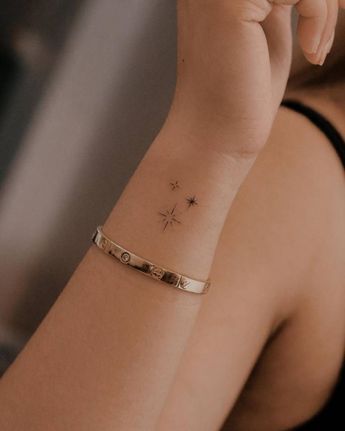Close-up of a delicate wrist tattoo featuring small stars and a compass, paired with a sleek gold bracelet.