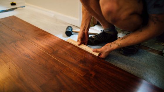 Person installing hardwood floor with a mallet and spacer on a carpeted surface.