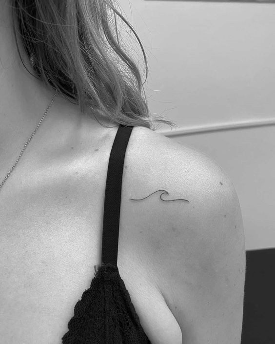 Minimalist wave tattoo on a woman's shoulder, black lace top visible, close-up shot in black and white.