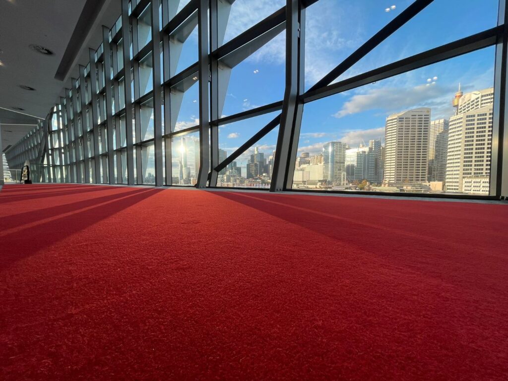 Modern building interior with red carpet and large windows overlooking a city skyline with skyscrapers and a blue sky.