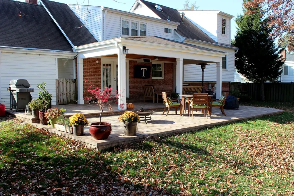 Spacious backyard patio with plants, outdoor furniture, and a grill. Covered seating area attached to a white brick house.