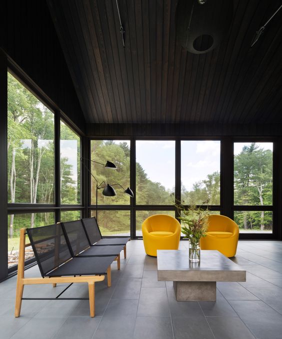 Modern living room with yellow chairs, large windows, black ceiling, minimalist furniture, and scenic forest view.