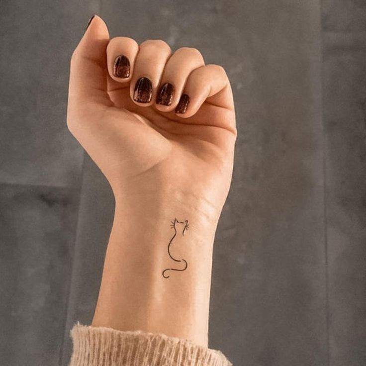 Minimalist cat tattoo on a wrist with dark nail polish. Simple black line art design on a shaded background.