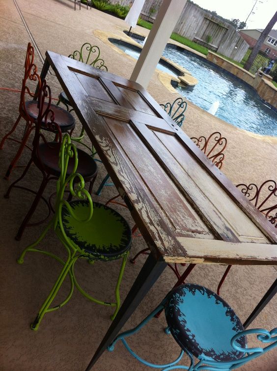 Vintage patio dining table made from an old wooden door paired with colorful, rustic metal chairs beside a pool.