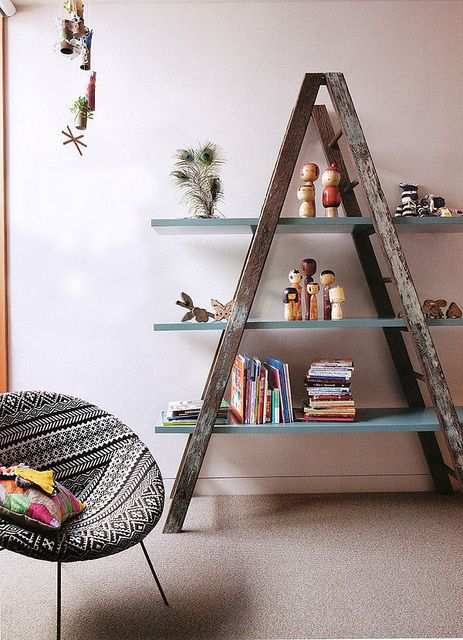 Stylish A-frame shelf with books, dolls, and trinkets beside a modern chair with black and white patterns in a cozy room.