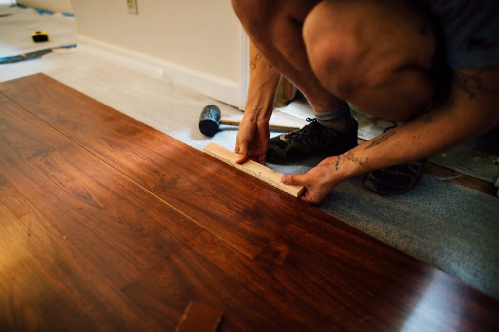 Person installing hardwood flooring with tools, close-up of hands working on the wooden floor during renovation.