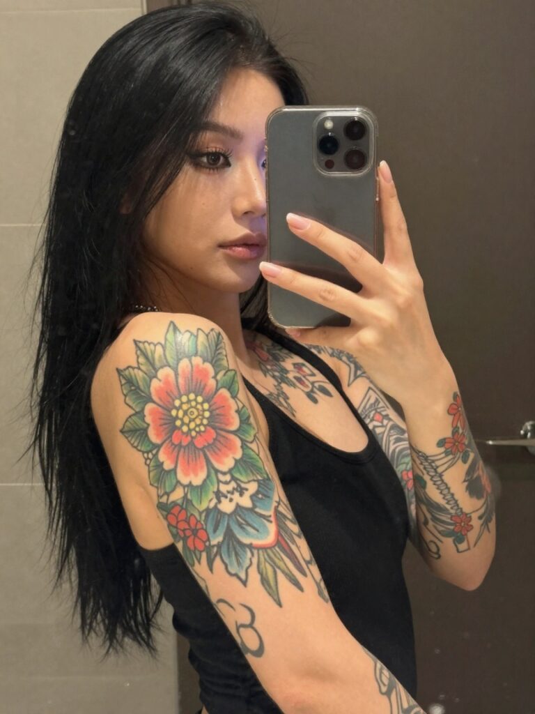 Woman taking a mirror selfie, showing colorful floral tattoos on her upper arm and shoulder while holding a phone.