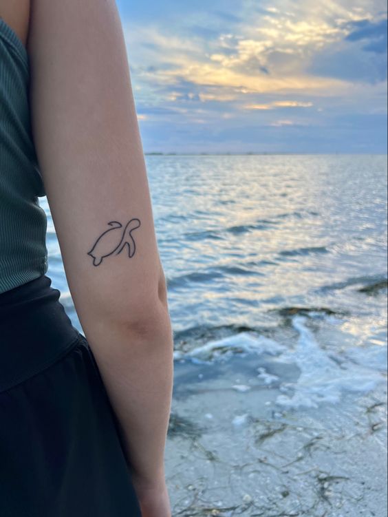 Person with a turtle tattoo on their arm standing by a serene beach at sunset.