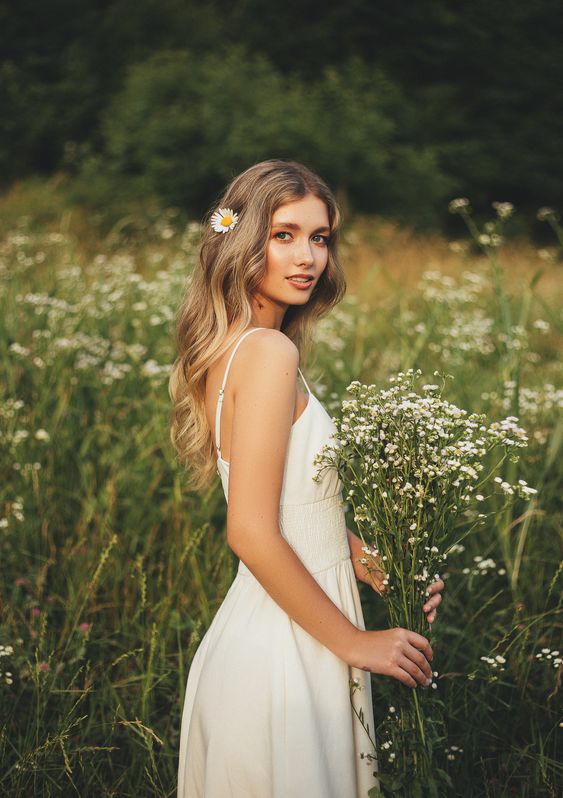 Woman in white dress holding a bouquet of wildflowers in a field, with a daisy in her hair, looking back.