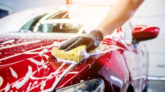 Hand washing a red car with a yellow sponge and soapy water to clean the hood, showcasing car care and maintenance.