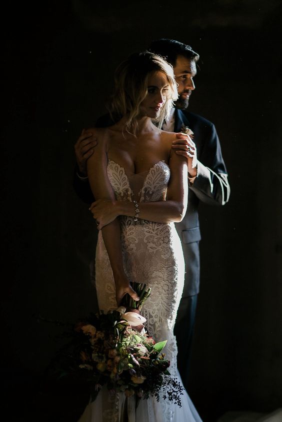 Bride and groom in dramatic lighting, embracing tenderly, bride holding bouquet, wearing lace wedding gown.