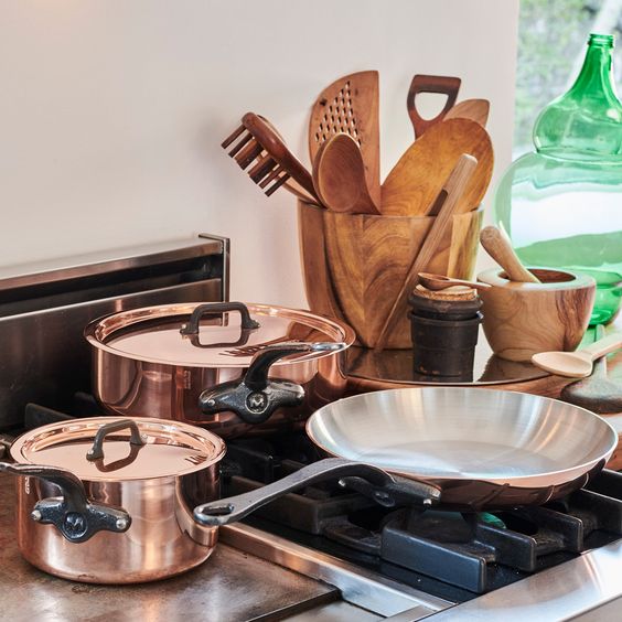 Copper cookware and wooden utensils on a gas stove in a modern kitchen. Rustic kitchenware and green glass vase nearby.