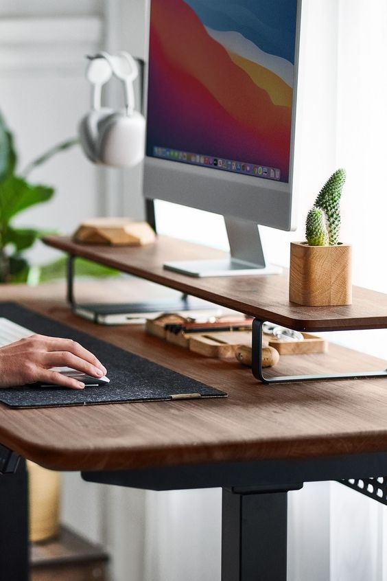 Modern home office setup with computer, headphones, cactus plant, and hand on mouse at wooden desk with natural light.