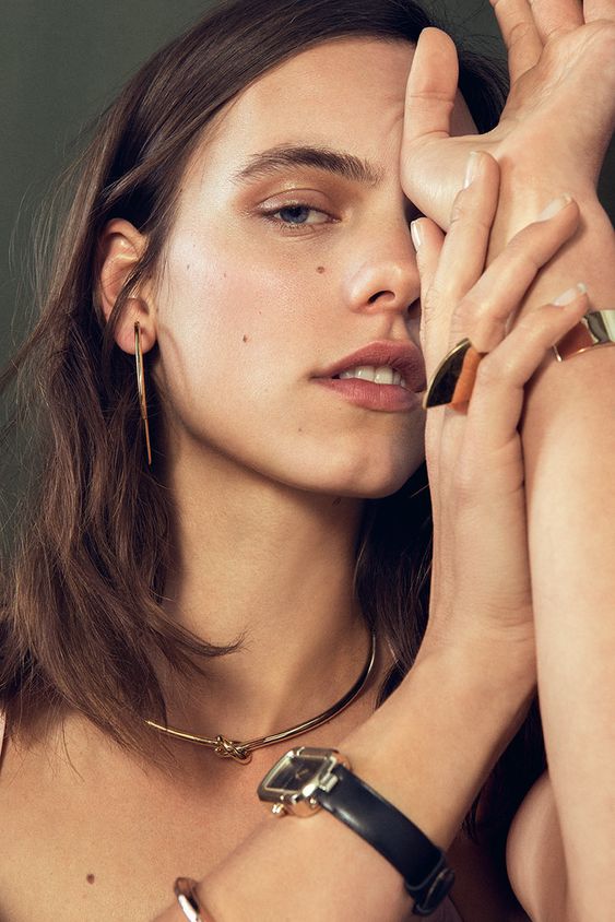 Woman with brown hair wearing gold jewelry and posing with hands, showcasing contemporary earrings, rings, and necklace.