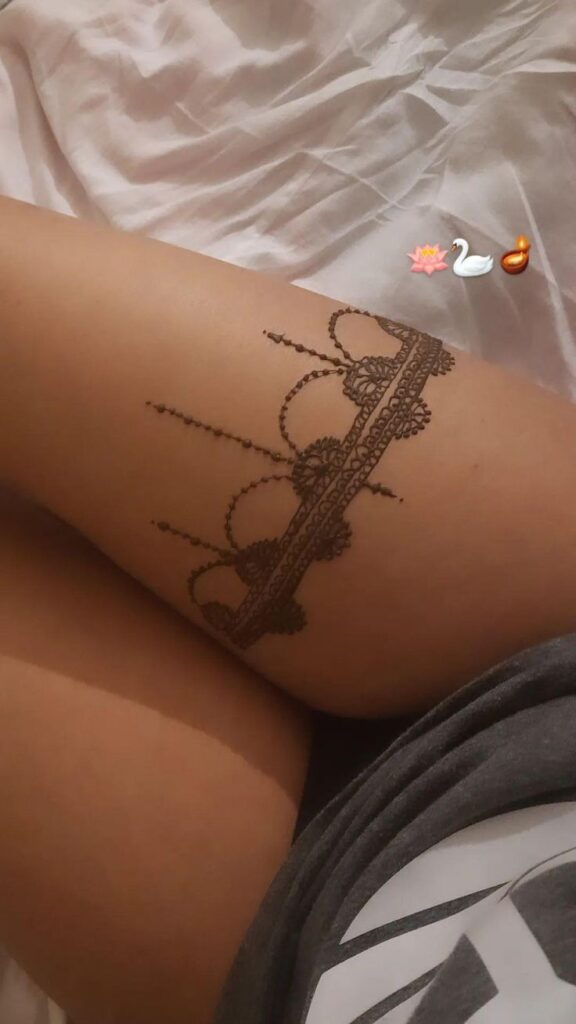 Ornate lace-patterned henna tattoo on thigh, shown against white bedding and grey clothing, featuring floral and swan emojis.