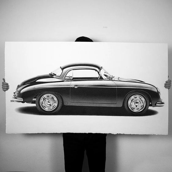 Person holding a large framed picture of a classic black sports car against a plain white background.