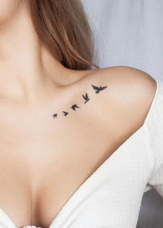 Minimalist bird tattoo on collarbone with five black birds in flight, worn by a woman in a white off-shoulder top.