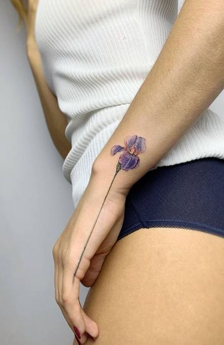 Purple flower tattoo on a woman's forearm, wearing a white ribbed tank top and dark blue shorts, minimalist design.