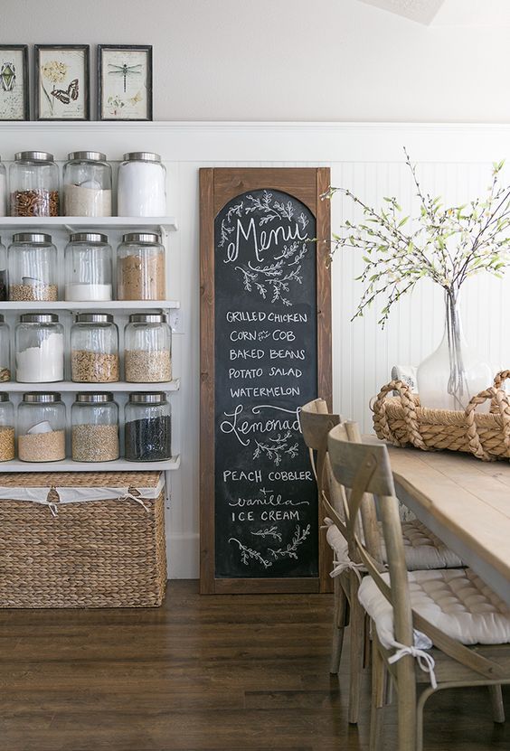 Rustic kitchen with a chalkboard menu, glass jars on open shelving, wicker basket, and a vase with branches on the dining table.