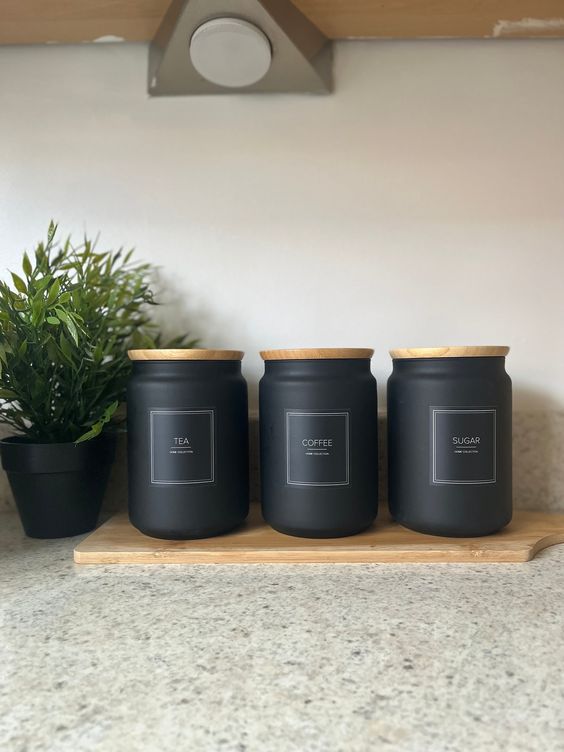 Black tea, coffee, and sugar canisters with wooden lids on a kitchen countertop, next to a small green plant.