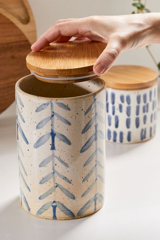 Hand lifting lid of a ceramic canister with blue leaf pattern, next to another canister on a kitchen counter.