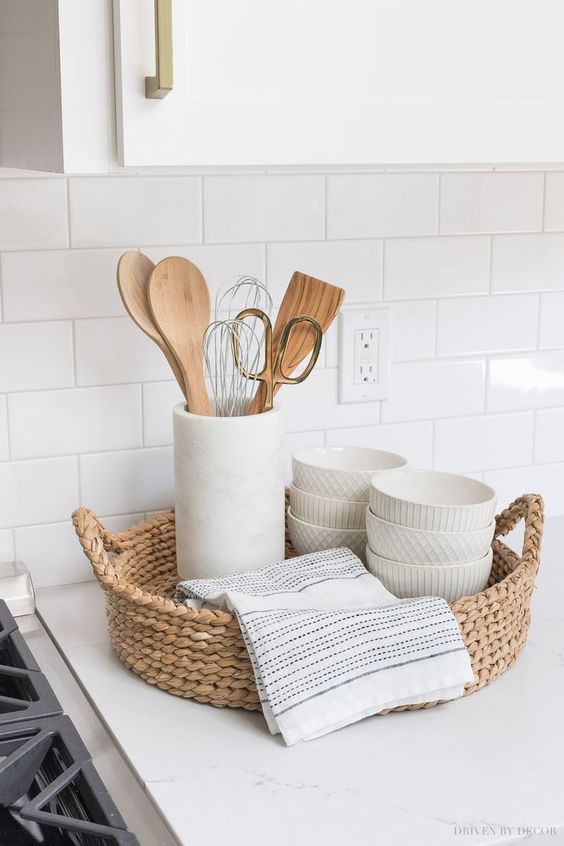 Modern kitchen with woven basket, white bowls, utensils, and towels on white countertop, against white subway tile backsplash.